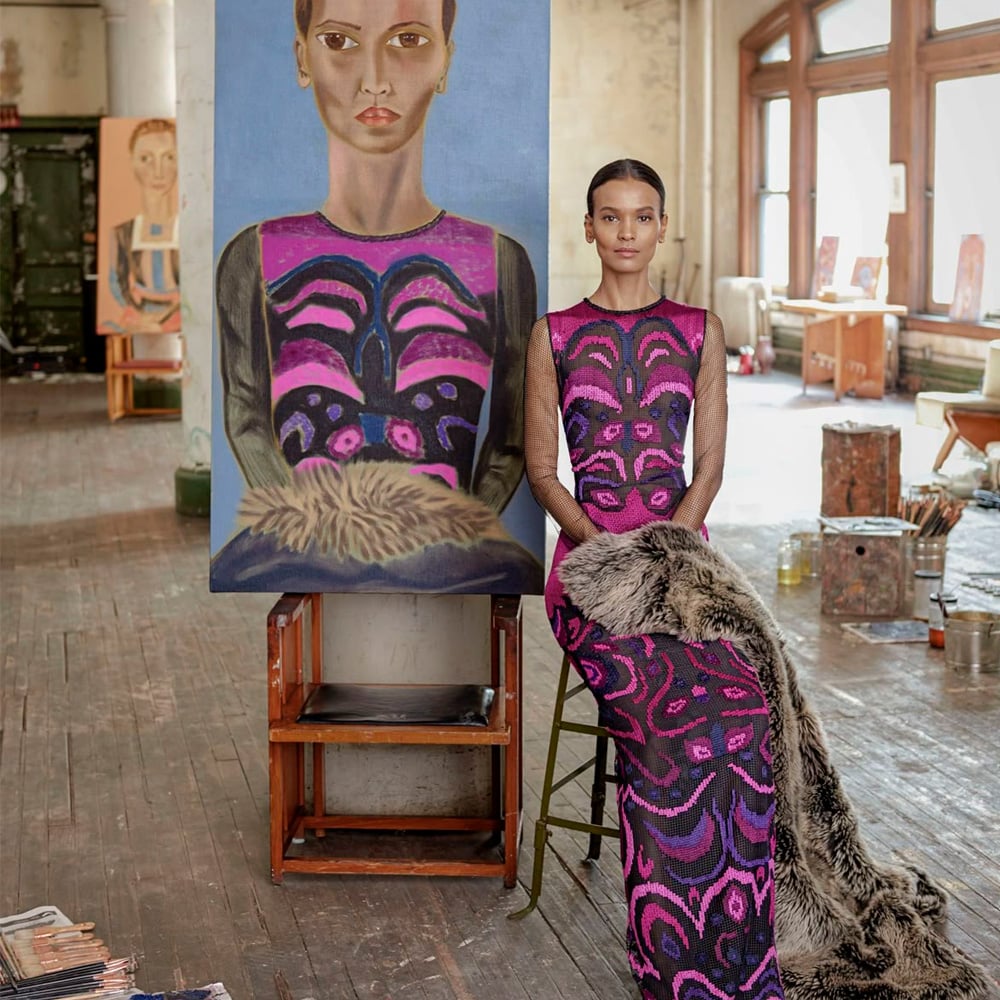 A woman in a vibrant, patterned dress and fur stole sits beside a large portrait that closely resembles her, inside an artist’s studio with wooden floors, art supplies, and paintings visible in the background.