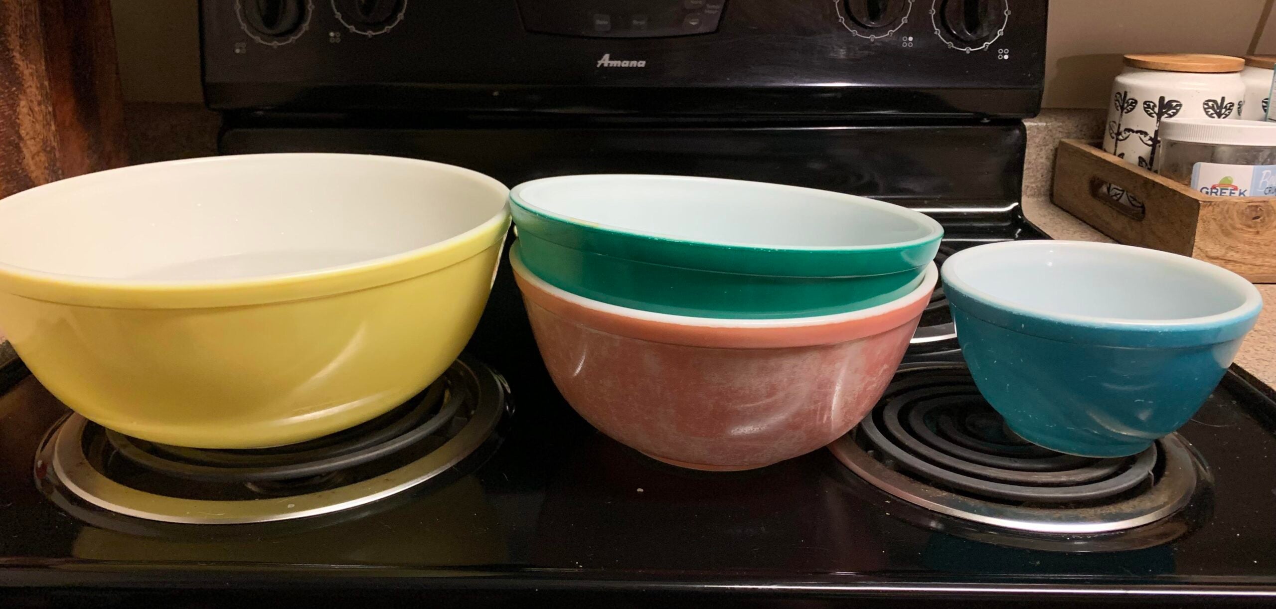 Four vintage mixing bowls in yellow, white, pink, green, and blue are nested and displayed on a black stovetop, with kitchen items visible in the background.