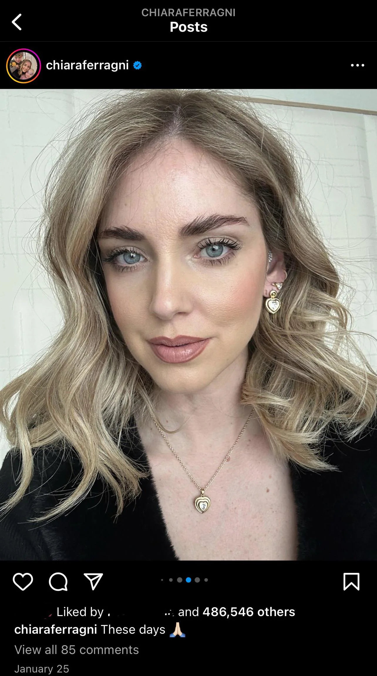 A woman with wavy blonde hair and blue eyes takes a close-up selfie. She is wearing a black top, layered gold necklaces, and heart-shaped earrings, with a soft, natural makeup look. The Instagram interface is visible.