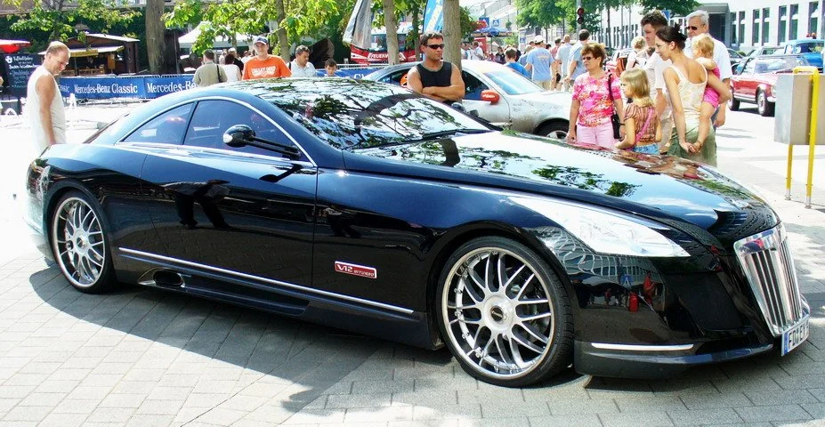A sleek, black luxury sports car is parked on a city street. Several people, including children, stand nearby admiring the vehicle. Trees and other cars are visible in the background on a sunny day.