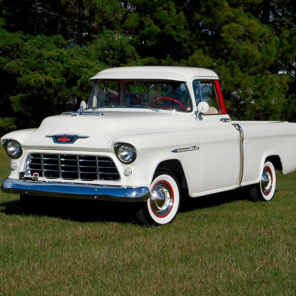 A vintage white Chevrolet pickup truck with red and chrome accents is parked on green grass, with trees in the background. The truck has classic round headlights and whitewall tires.