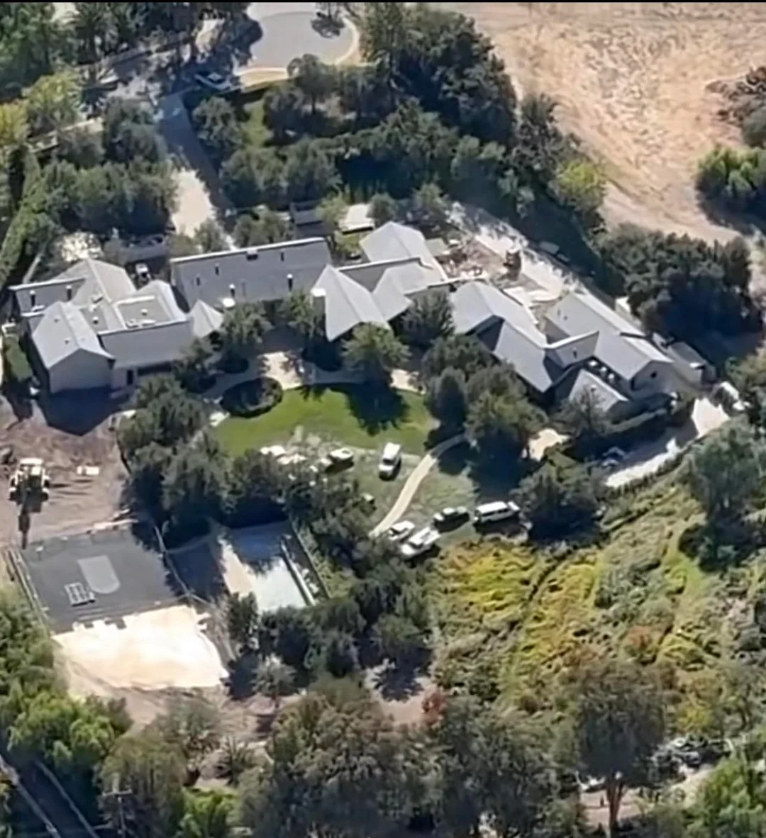 Aerial view of a large estate with multiple buildings, a driveway, green lawns, trees, and recreational areas including a tennis or basketball court, surrounded by natural landscape and vegetation.