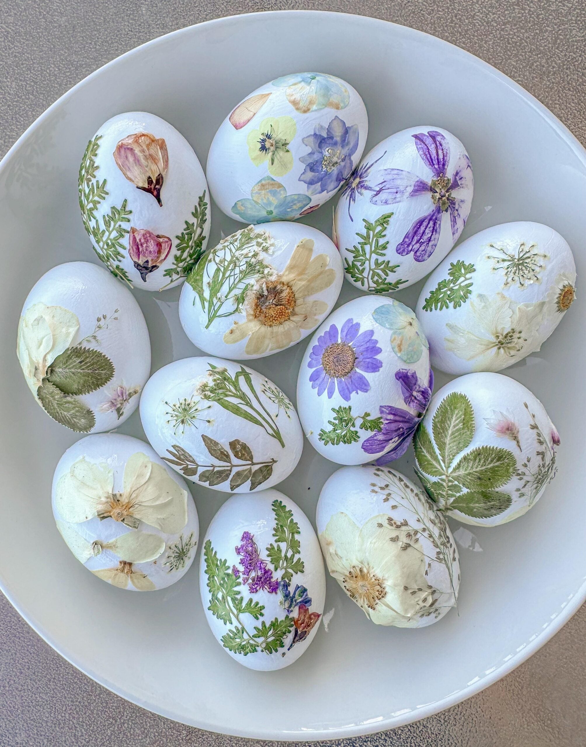 A white bowl filled with eggs decorated with colorful pressed flowers, leaves, and botanical designs, creating a delicate and artistic spring or Easter theme.