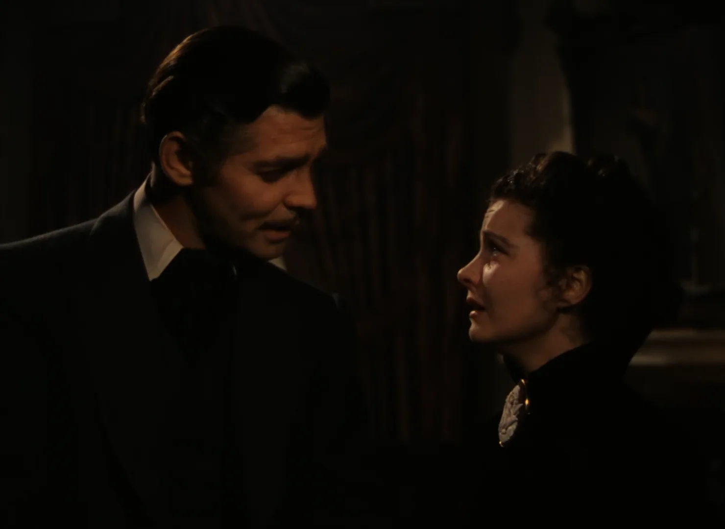 A man in a dark suit and a woman in a black dress with an ornate collar stand close together indoors, facing each other and appearing to have an emotional conversation in low lighting.