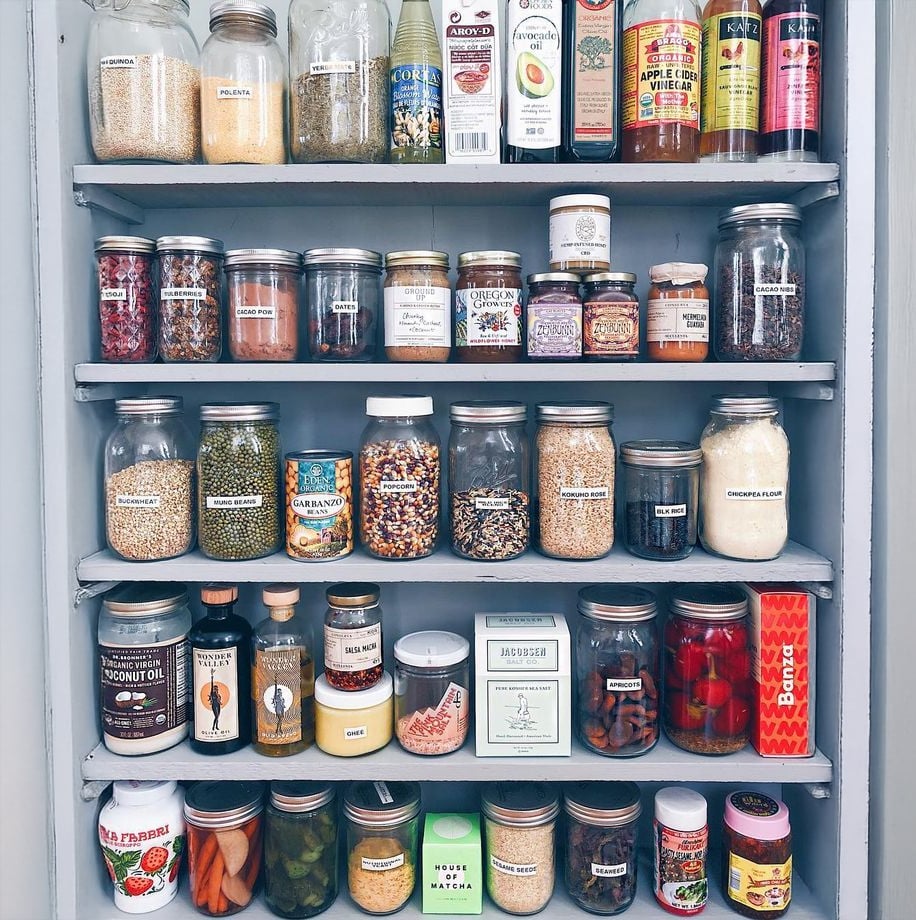 A neatly organized pantry shelf holds various jars and cans of grains, spices, oils, vinegars, and other preserved foods, all labeled and arranged by size, creating a colorful and orderly display.