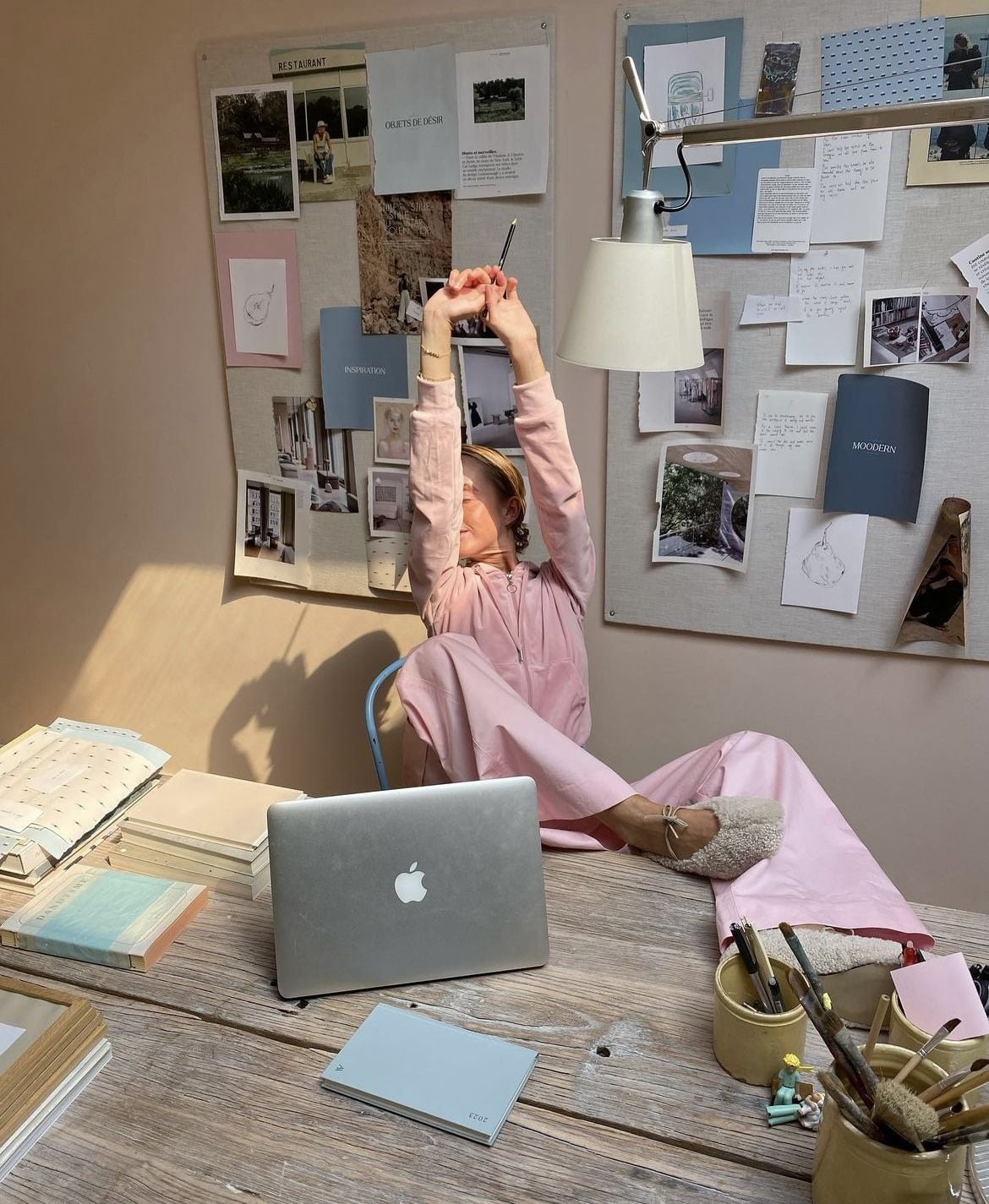 A person in pink loungewear stretches at a desk with a laptop, notebooks, and stationery. Sunlight shines on them. Behind, wall boards display photos, papers, and notes, creating a cozy, creative workspace.