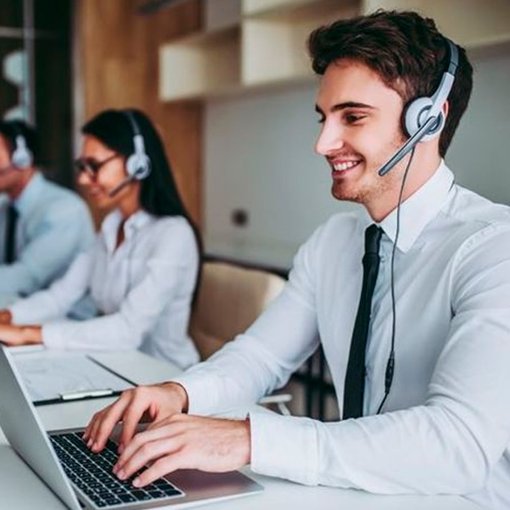 A young man wearing a headset and white shirt smiles while working on a laptop in an office. Two colleagues, also wearing headsets, work at desks beside him.