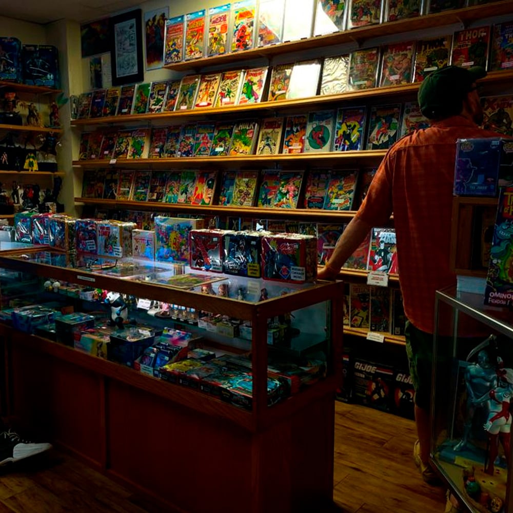 A man in a cap and orange shirt browses a comic book store filled with colorful comic books displayed on shelves and in glass cases, with action figures also visible on display.