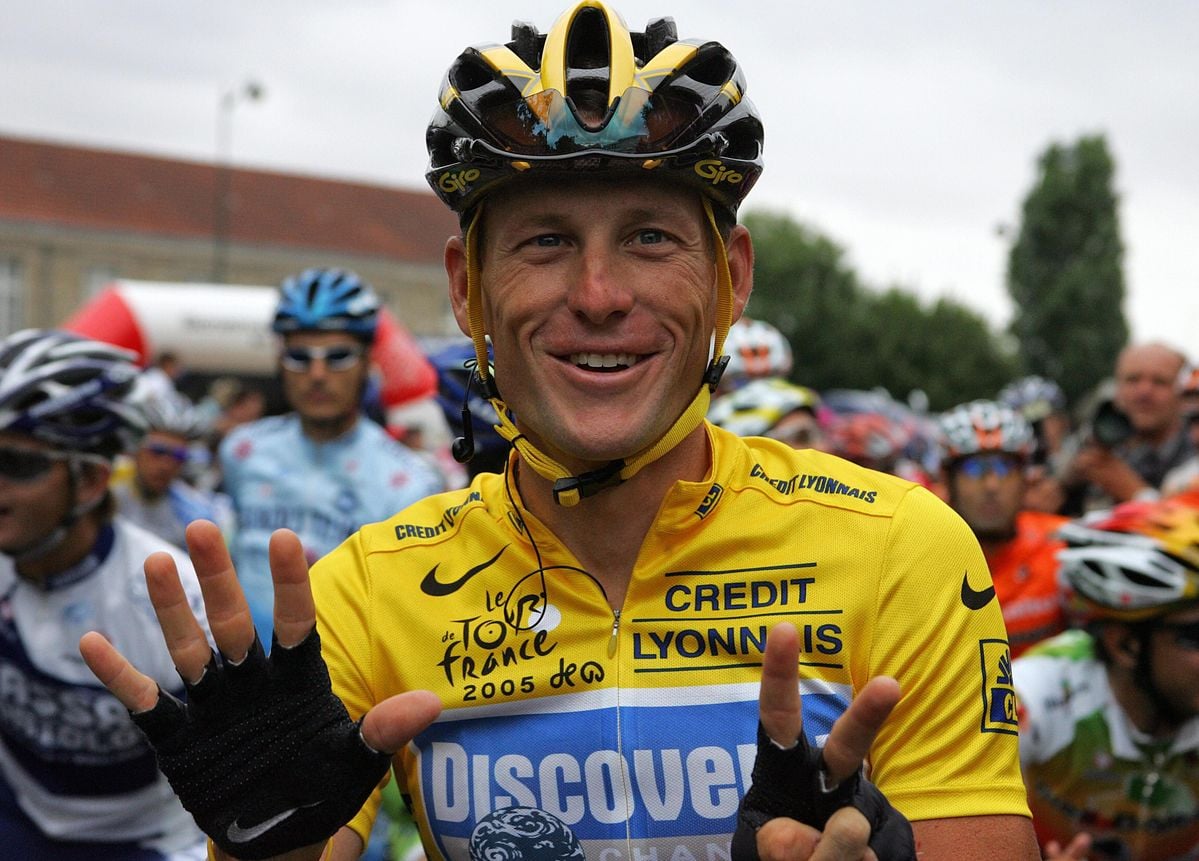 A smiling cyclist in a yellow jersey and helmet holds up seven fingers at a race start, surrounded by other riders. His jersey reads "Discovery Channel" and "Credit Lyonnais," with "Tour de France 2005" written on it.