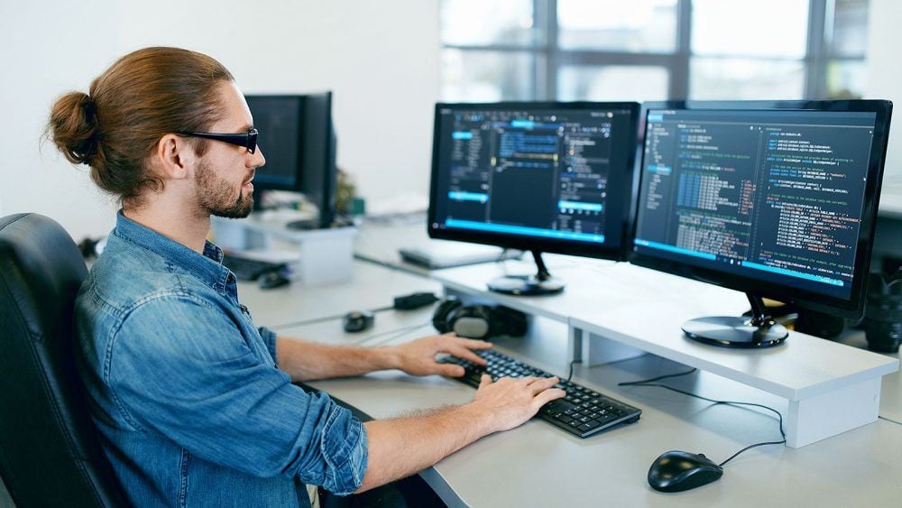 A man with glasses and a ponytail sits at a desk typing on a keyboard, facing two monitors displaying code in a modern, bright office environment.