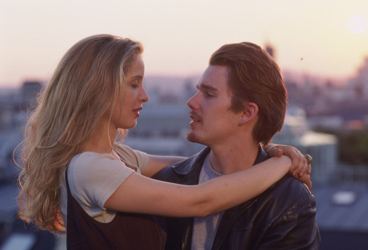 A woman with long blonde hair embraces a man with short brown hair and a goatee. They gaze into each other's eyes on a rooftop at sunset, with a blurred city skyline in the background.