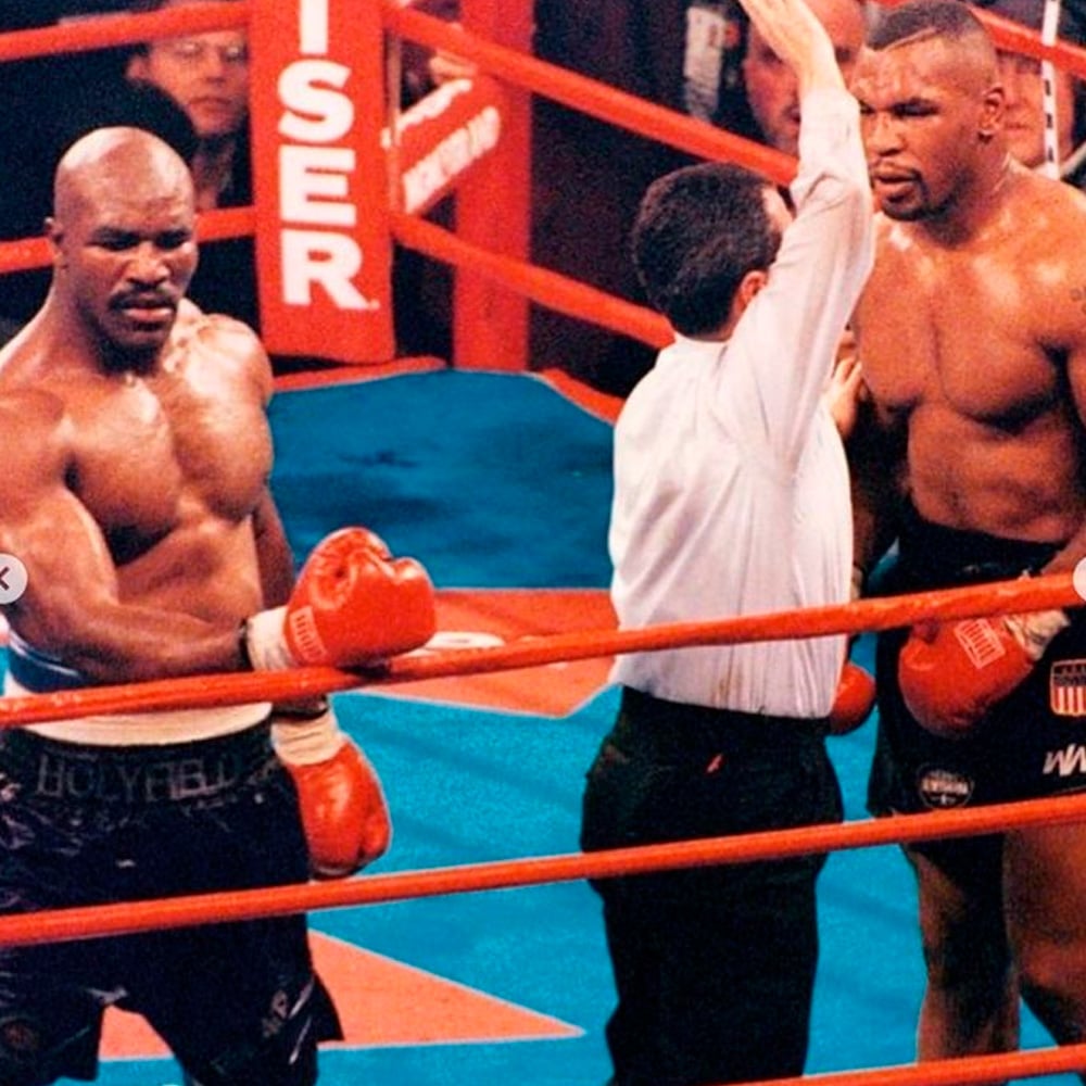Two male boxers stand in a boxing ring with a referee between them. One boxer is wearing red gloves and black shorts labeled "Holyfield," while the other is in black shorts. The referee is raising his arm to signal a decision.