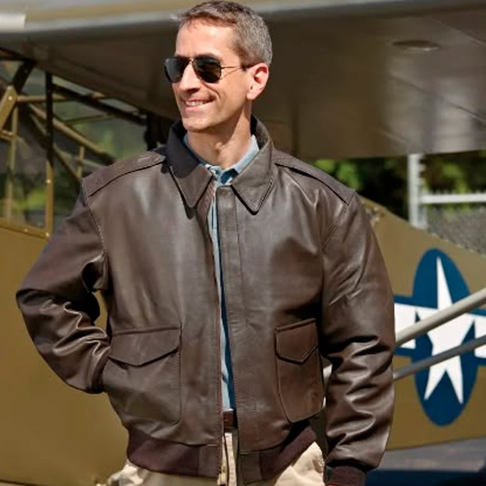 A man wearing sunglasses, a brown leather bomber jacket, and khaki pants stands in front of a vintage military airplane with a star insignia, smiling and looking to the side.