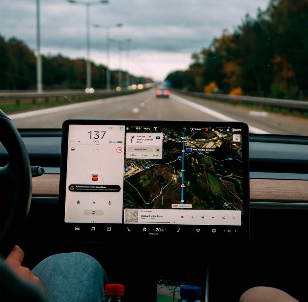 A car is being driven on a highway, showing a dashboard with a digital screen displaying speed, navigation, and music controls. The view outside includes a tree-lined road and a few cars ahead.