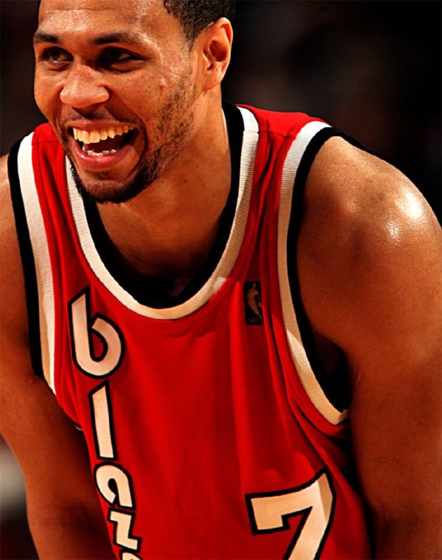 A basketball player in a red Portland Trail Blazers jersey with the number 7 smiles during a game.
