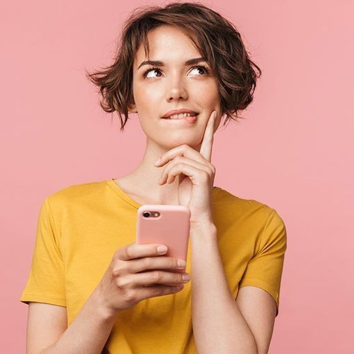 A young woman with short brown hair wearing a yellow shirt holds a pink smartphone and looks thoughtfully upward, with her finger resting on her cheek, against a pink background.