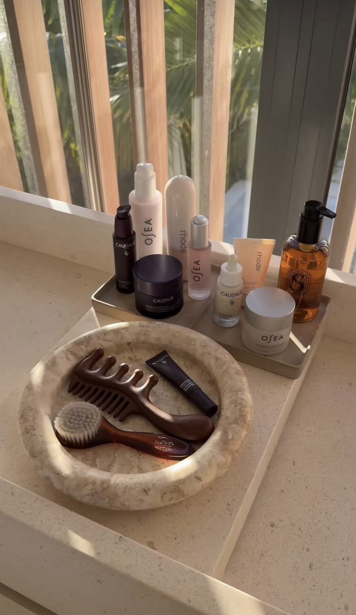 A stone dish holding a wooden comb, a face brush, and a dark tube sits on a counter next to neatly arranged skincare bottles and jars, with sunlight streaming through window blinds behind them.