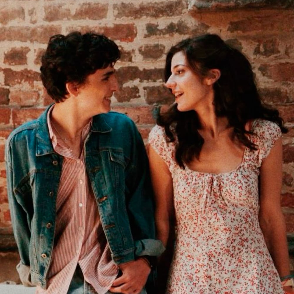Two young people, one wearing a denim jacket and the other in a floral dress, smile at each other while walking together in front of a brick wall.