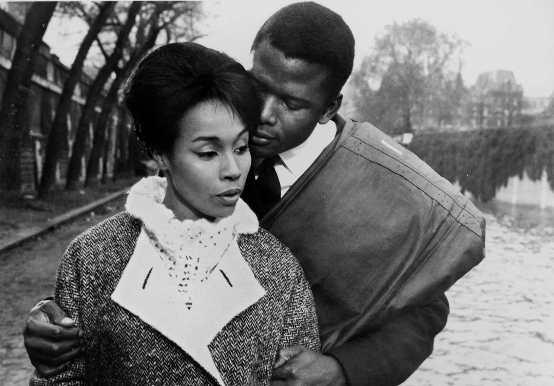 A Black man embraces a Black woman from behind as they stand near a river on a tree-lined path. The woman looks thoughtfully ahead while the man gazes at her. Both wear coats and it appears to be a cool day. The image is in black and white.