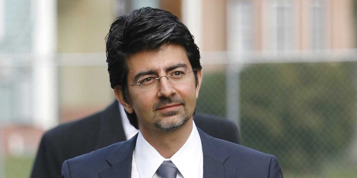 A man with dark hair, glasses, and a goatee wearing a dark suit, white shirt, and blue tie stands outdoors, with a blurred background featuring a fence and buildings.