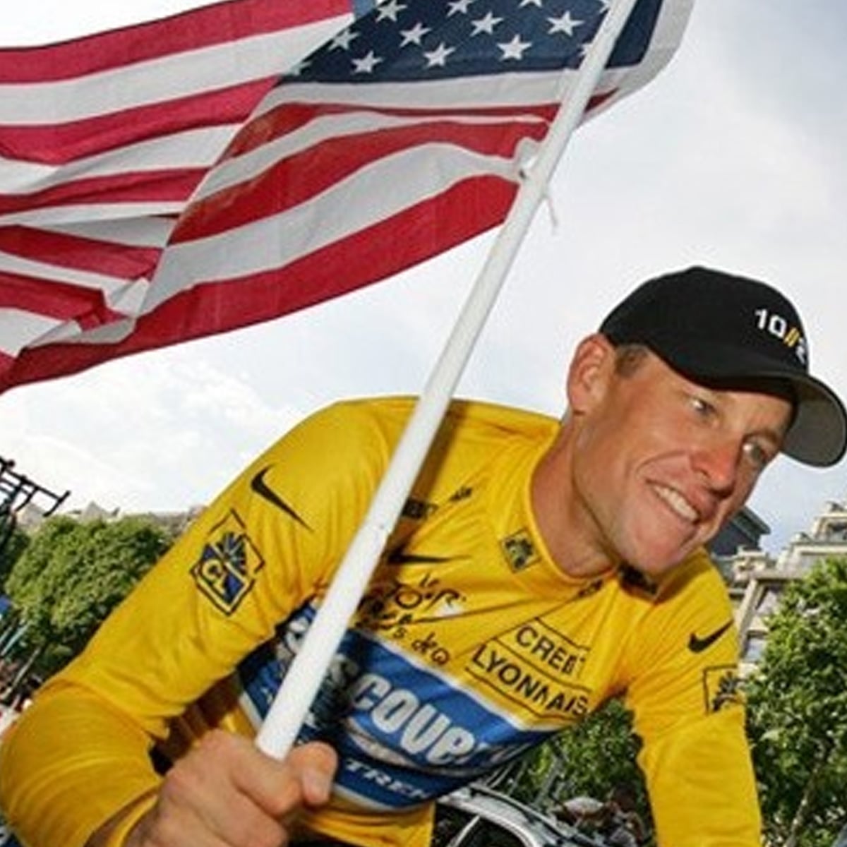 A cyclist in a yellow jersey and black cap smiles while holding a large American flag. Trees and parts of a street are visible in the background.