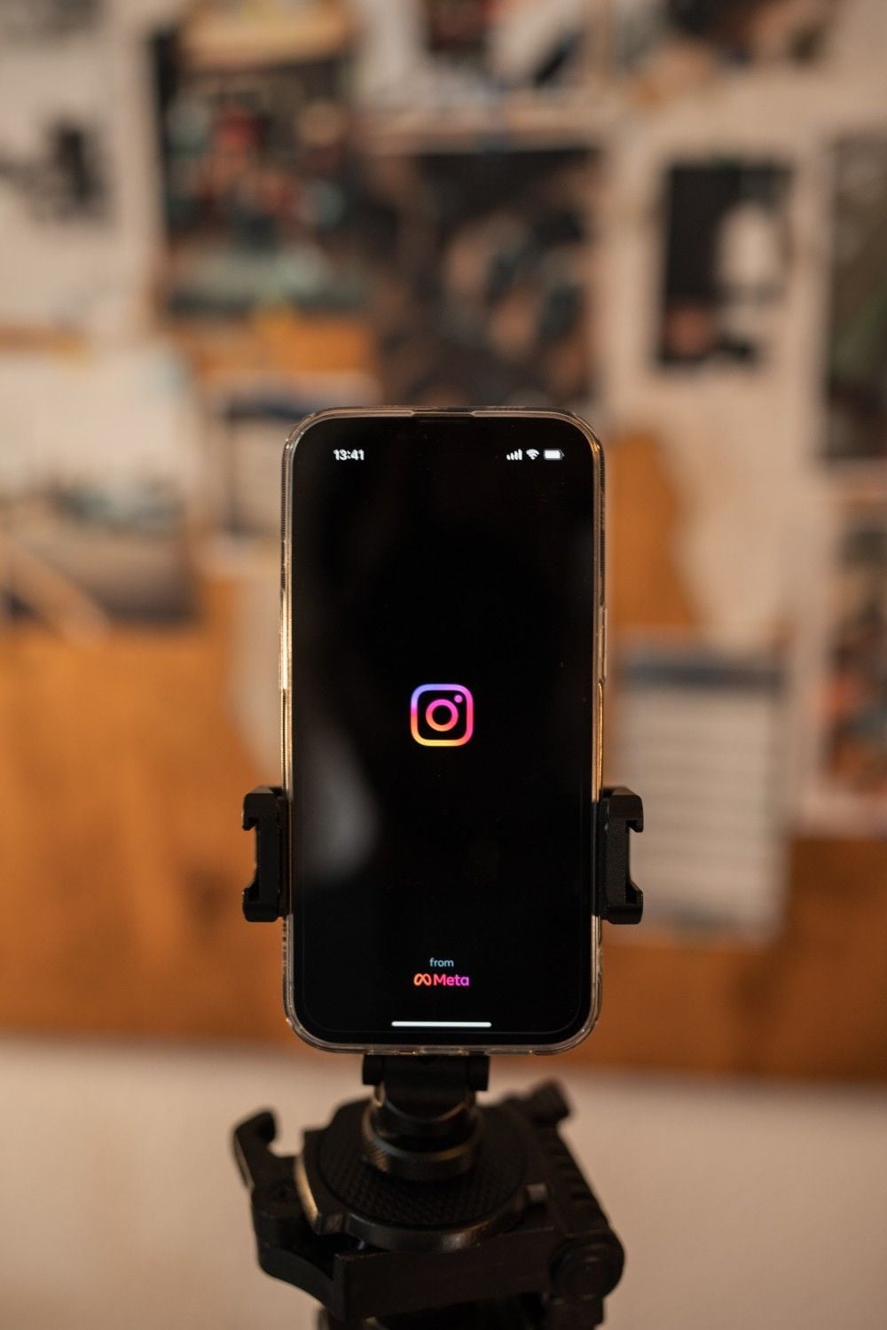 A smartphone mounted on a tripod displays the Instagram logo on its screen, with a blurred corkboard and photos in the background.