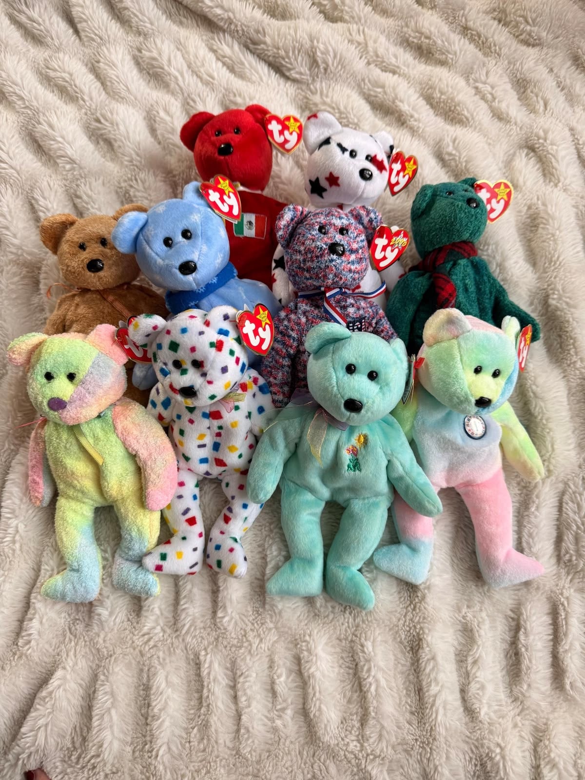 A collection of ten colorful TY Beanie Baby teddy bears with heart-shaped tags, arranged on a textured beige blanket. The bears come in various colors and patterns, including pastel, solid, and speckled designs.
