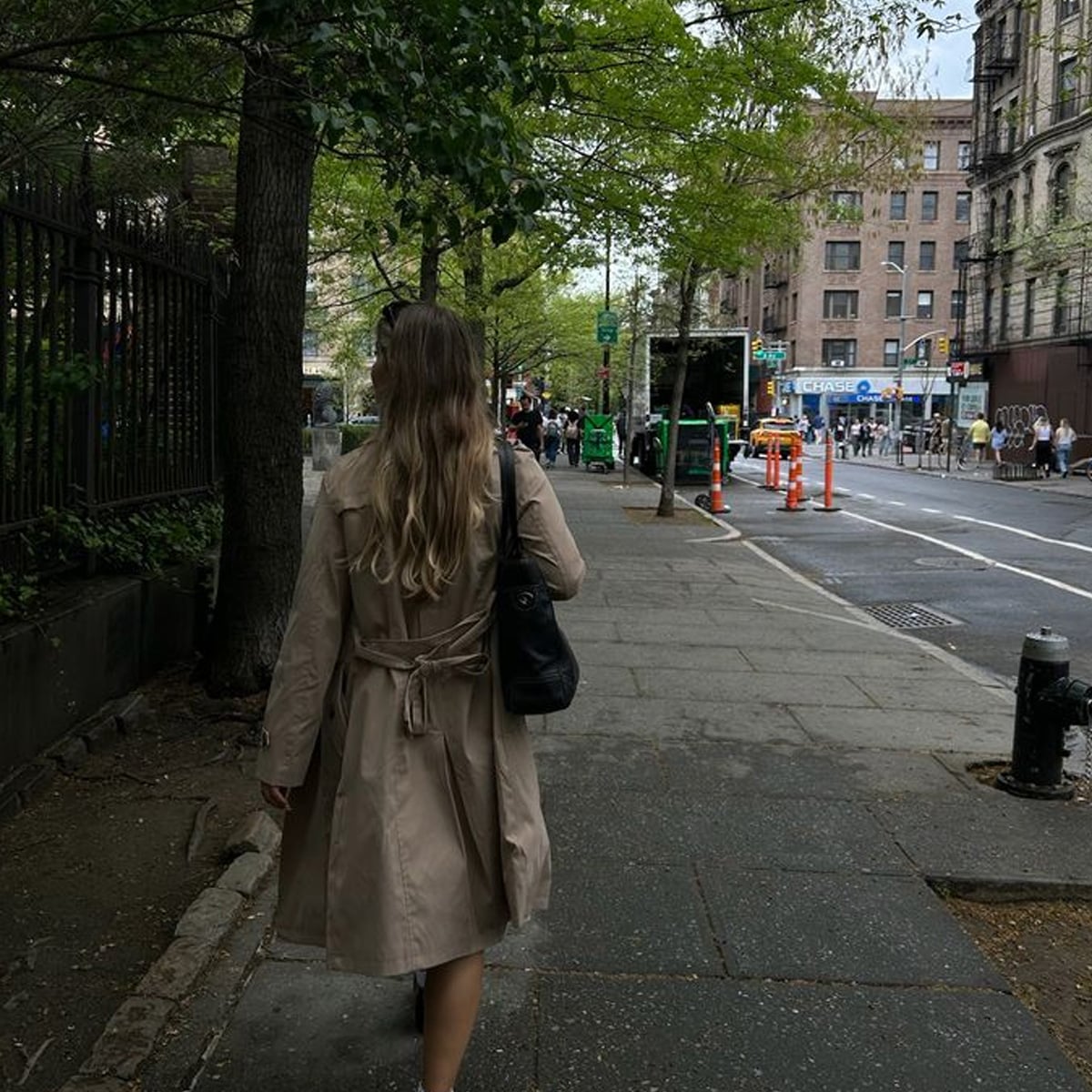 A woman with long hair wearing a beige trench coat walks along a city sidewalk bordered by trees and buildings, while cars and people are visible in the background on a cloudy day.