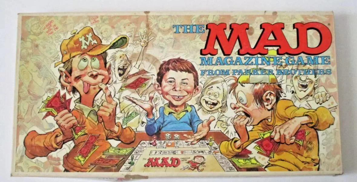 Cartoon illustration of four goofy, excited people playing The MAD Magazine Game board game. One character wears an "M" cap, and Alfred E. Neuman smiles widely in the center. The game box is colorful and humorous.