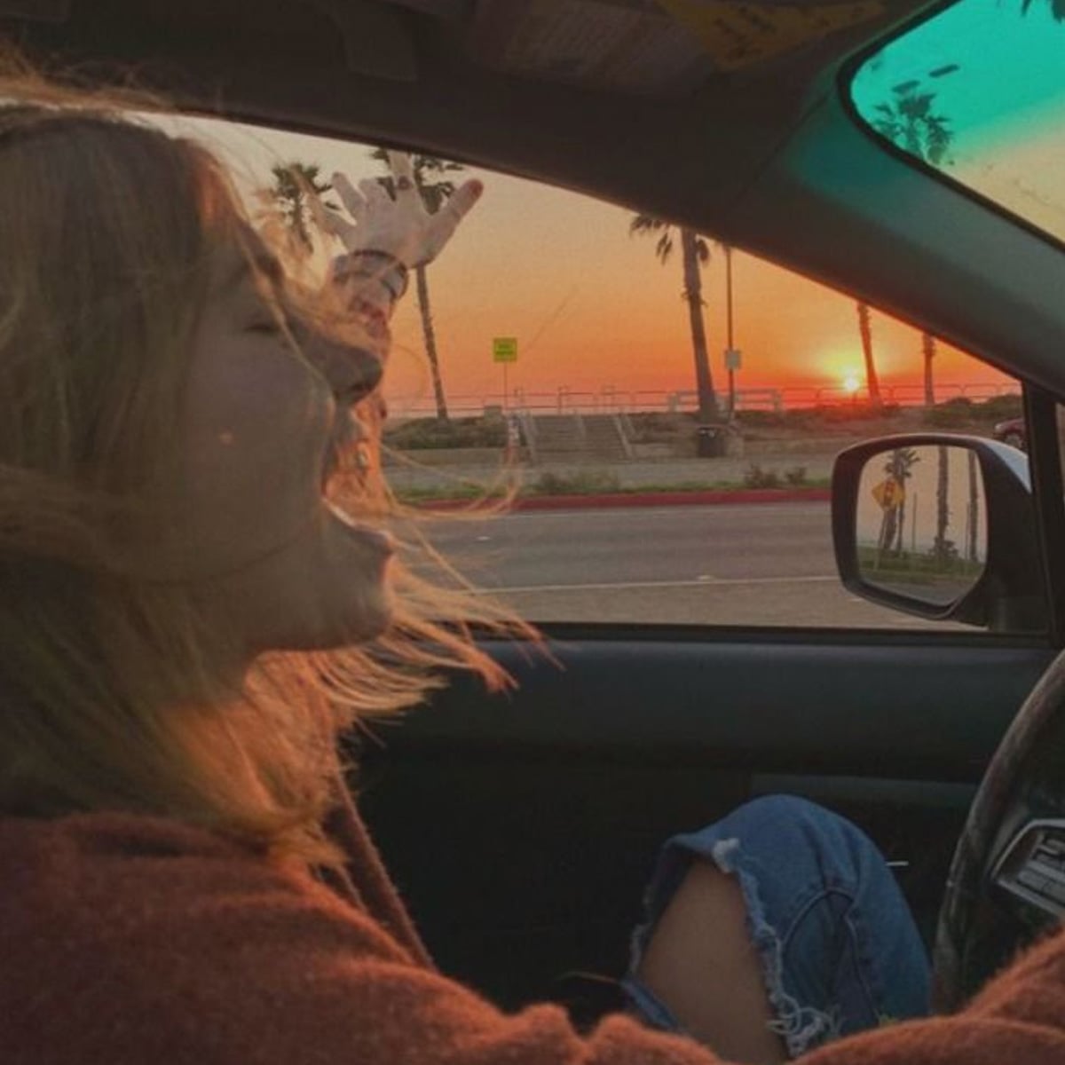 A person with shoulder-length hair sits in a car, smiling joyfully with one arm raised, enjoying a colorful sunset by the ocean with palm trees visible outside the window.