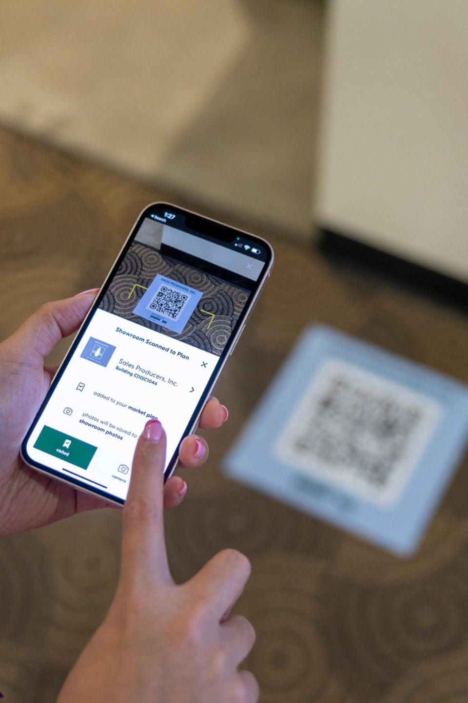 A person uses a smartphone to scan a QR code on the floor. The phone screen shows a payment interface with options, and the person is tapping a button with their index finger.