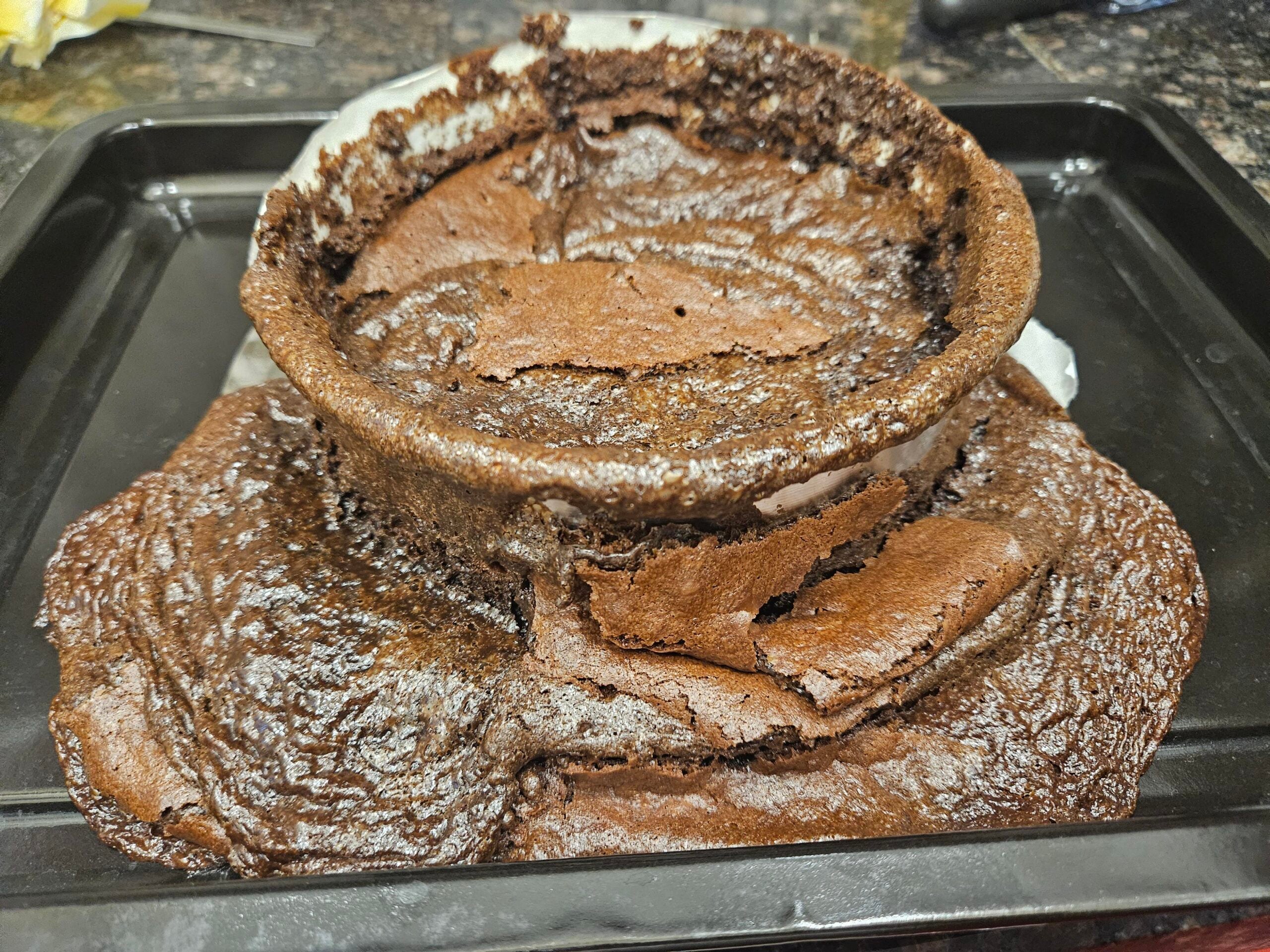 A failed chocolate cake sits on a black baking tray. The cake has collapsed in the center and the sides have overflowed, creating a sunken, uneven, and cracked appearance with unevenly baked edges.