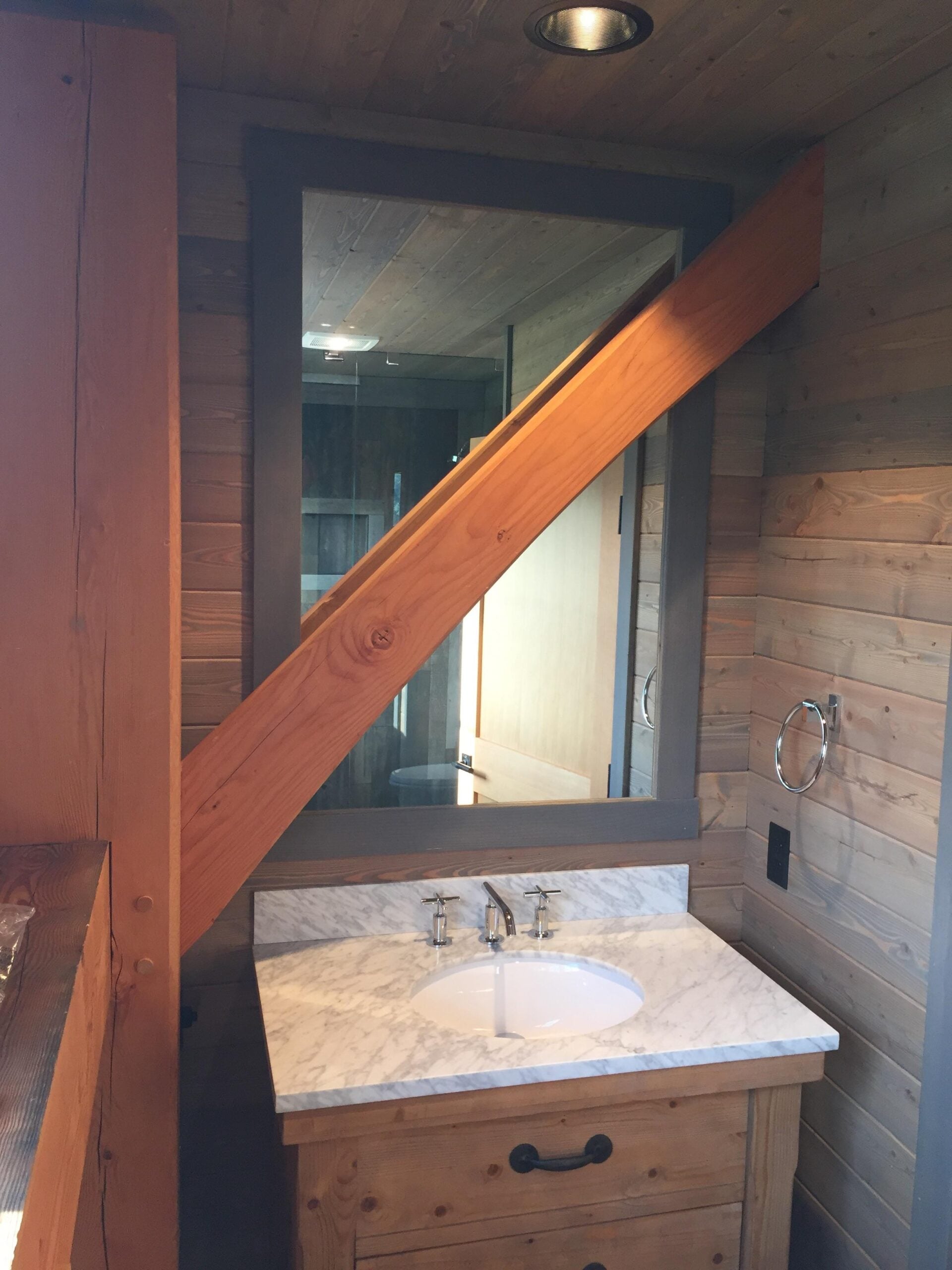 A rustic bathroom with wood-paneled walls, a marble countertop with a sink, and a large mirror partially obstructed by an angled wooden beam. A towel ring and electrical outlet are mounted on the wall.