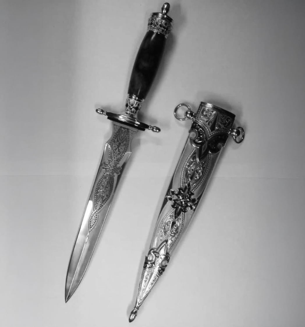 An ornate dagger with an engraved blade and elaborate handle lies next to its matching, intricately decorated sheath on a plain background. The image is in black and white.