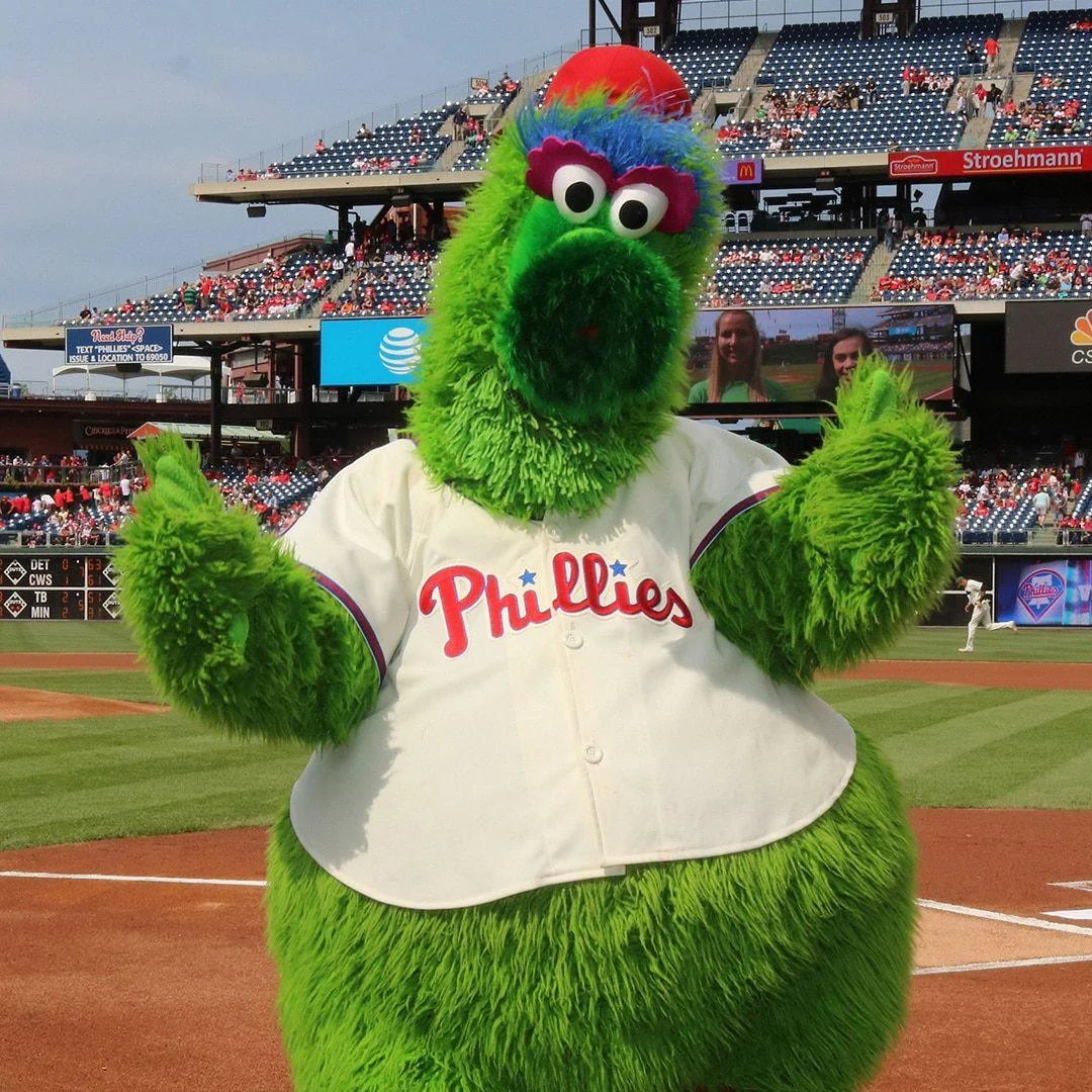 The Phillie Phanatic, a large green furry mascot in a Phillies jersey, stands on a baseball field with arms raised, with stadium seats and fans in the background.