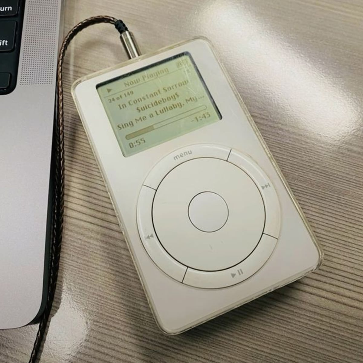 A first-generation Apple iPod with a white body and physical scroll wheel displays a song playing on its monochrome screen. It's connected to a braided headphone cable and resting on a wooden desk beside a laptop.