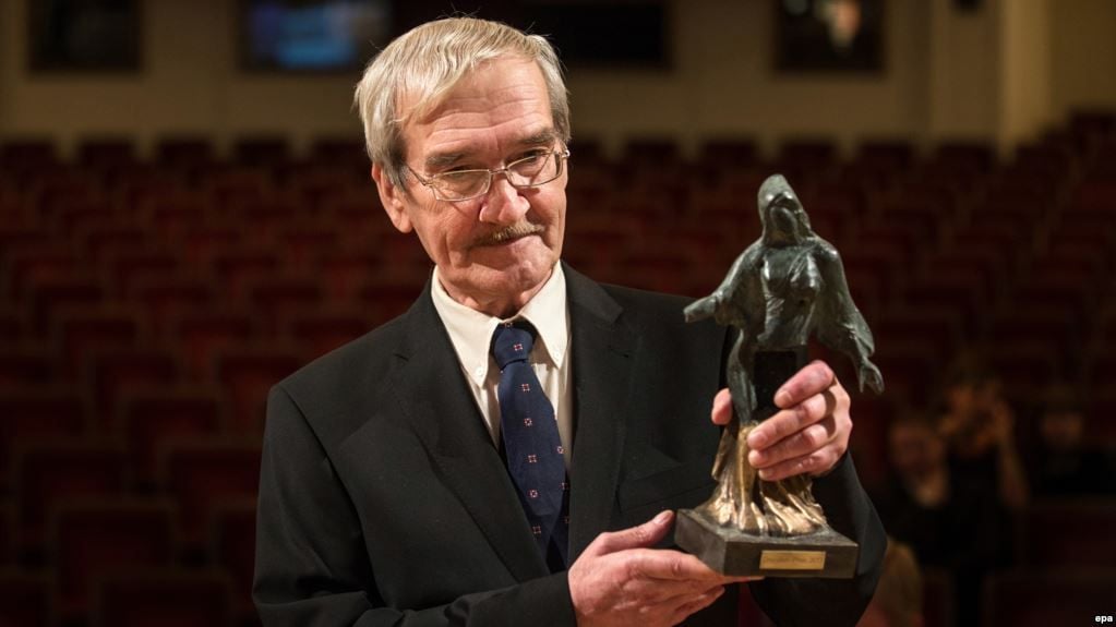 An older man in a suit and tie stands in an auditorium holding a statuette shaped like a cloaked figure, with empty red seats visible in the background.