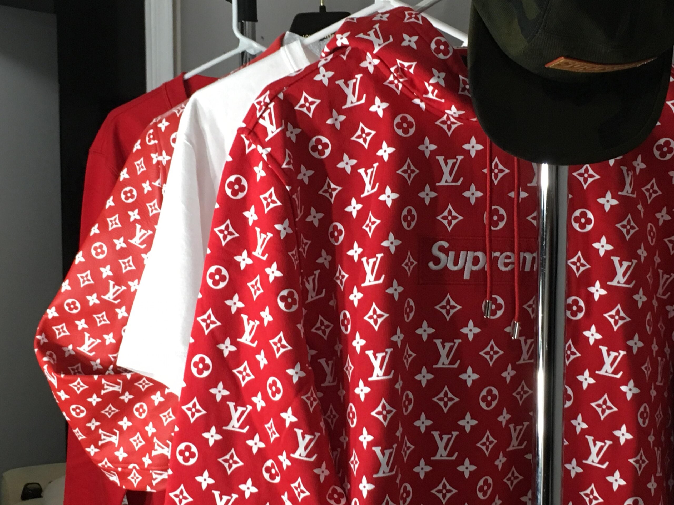 Three clothing items hang on hangers, including a red and white hoodie with a Louis Vuitton and Supreme logo pattern, a red shirt, and a white shirt. A green cap with an orange tag sits on top of the hangers.