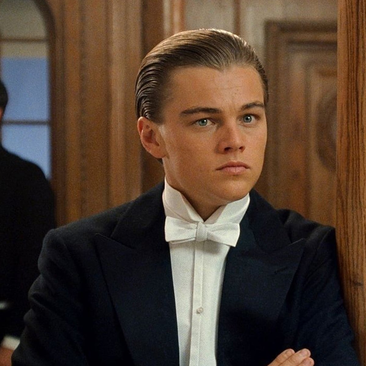 A young man with slicked-back hair, wearing a black tuxedo and white bow tie, stands indoors with a serious expression, leaning against a wooden surface.