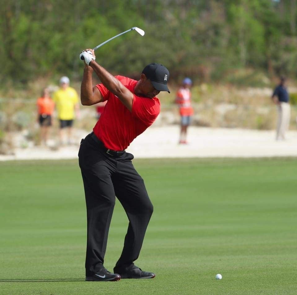 A golfer in a red shirt and black pants prepares to swing at a golf ball on a green course, with several people and trees blurred in the background.