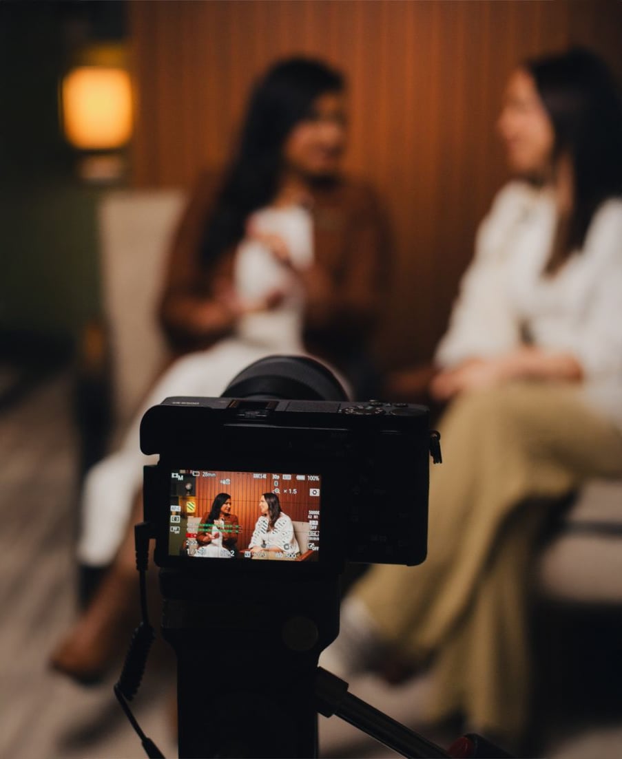 A camera films two women having a conversation while sitting together indoors. The women are out of focus in the background, but appear clearly on the camera’s display screen in the foreground.