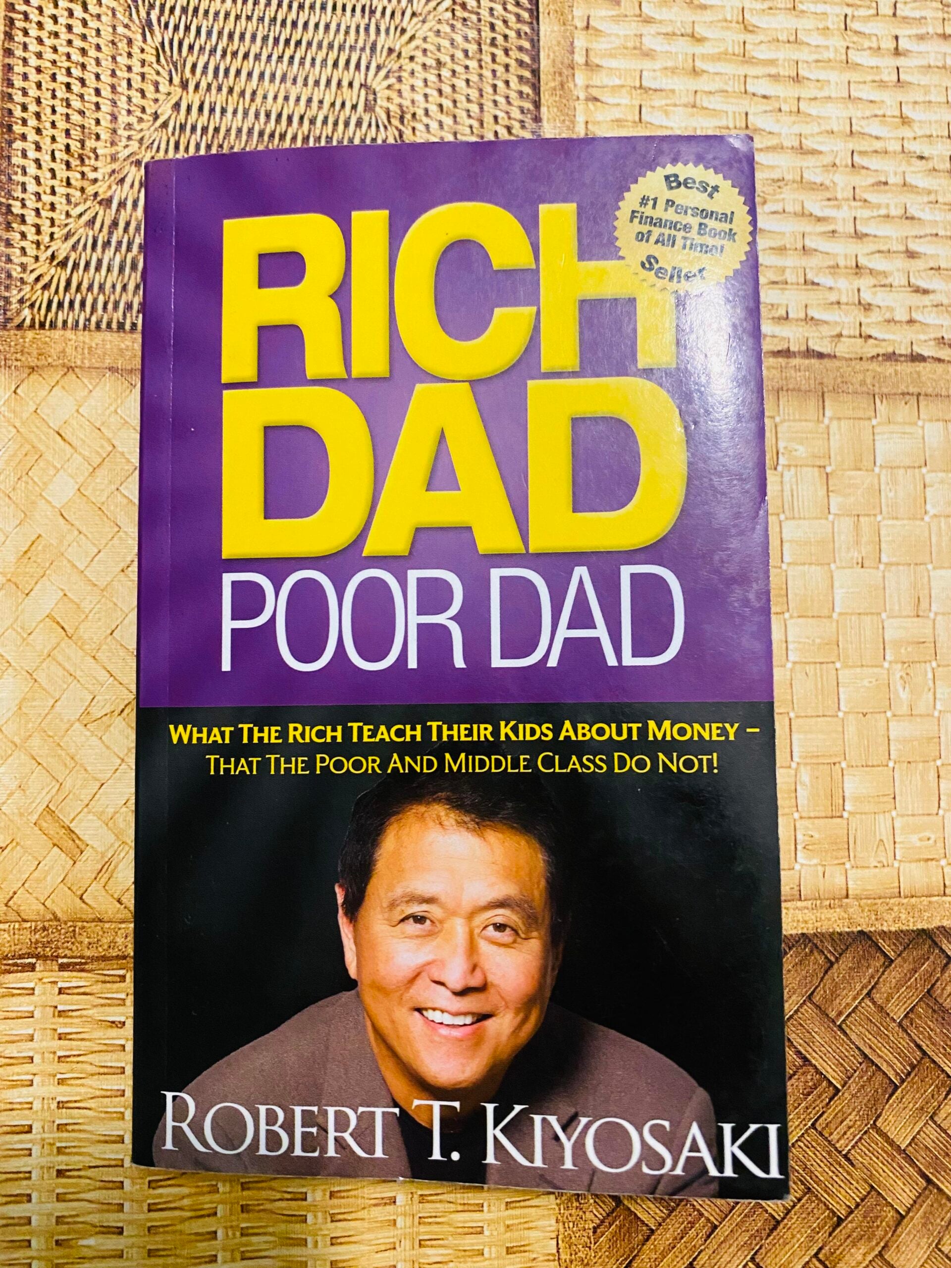 The book "Rich Dad Poor Dad" by Robert T. Kiyosaki is placed on a woven mat. The cover is purple and yellow, featuring a photo of the author and the subtitle about lessons on money.