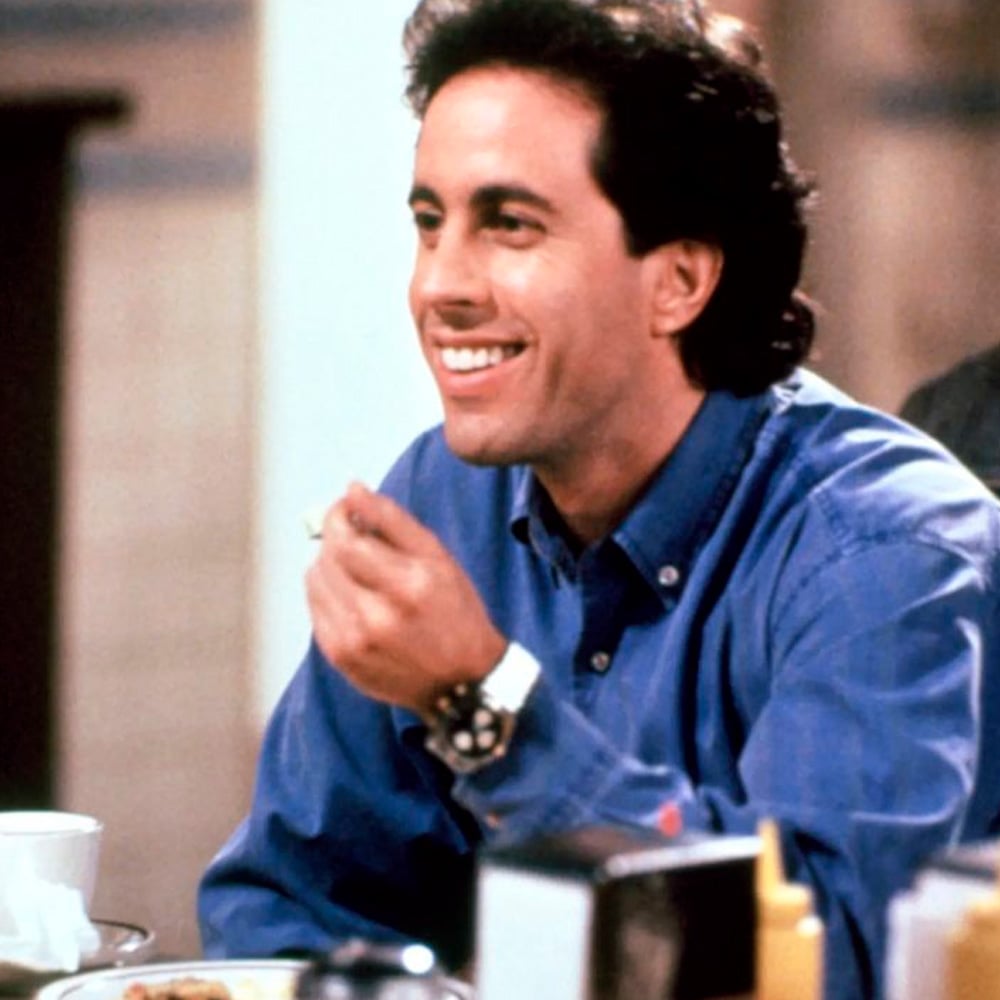 A man with dark curly hair wearing a blue shirt smiles while holding something in his hand, seated at a diner table with a coffee cup, condiments, and napkin holder in front of him.