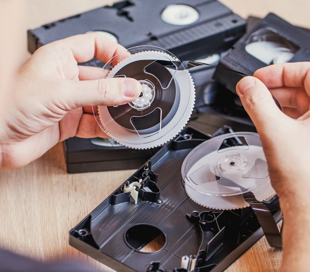 A person repairs a VHS tape, holding the reel and magnetic tape, with a disassembled cassette and other VHS tapes on a wooden surface.