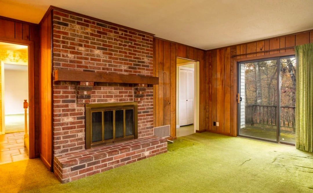 A retro living room with wood-paneled walls, green carpet, a brick fireplace, sliding glass door with green curtains, and two doorways leading to other rooms.