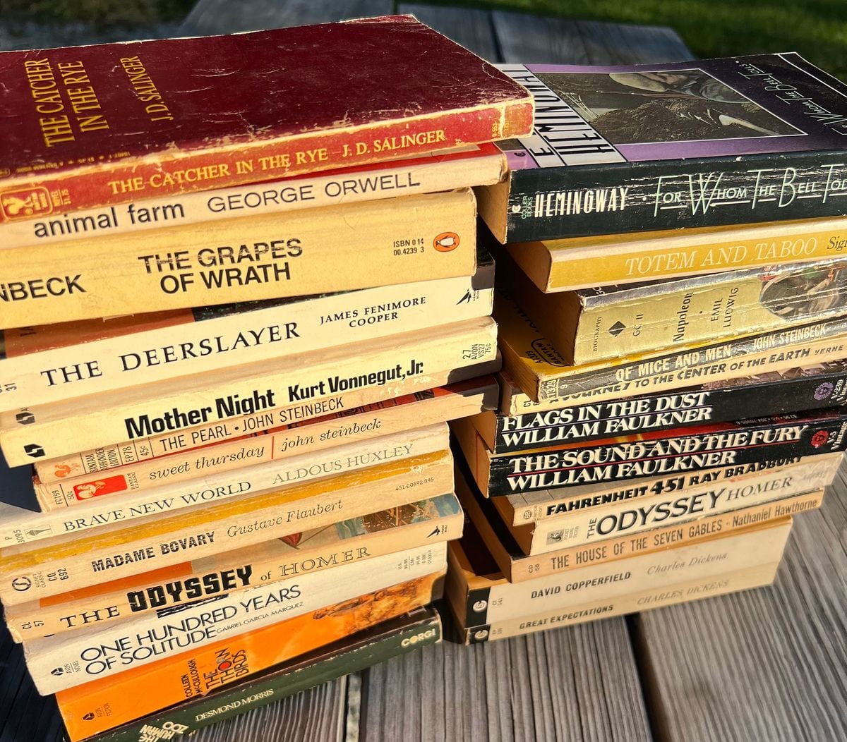 Two stacks of classic novels with yellowed covers, including titles like "The Catcher in the Rye," "Animal Farm," "The Grapes of Wrath," "Fahrenheit 451," and "One Hundred Years of Solitude," on a wooden outdoor table.
