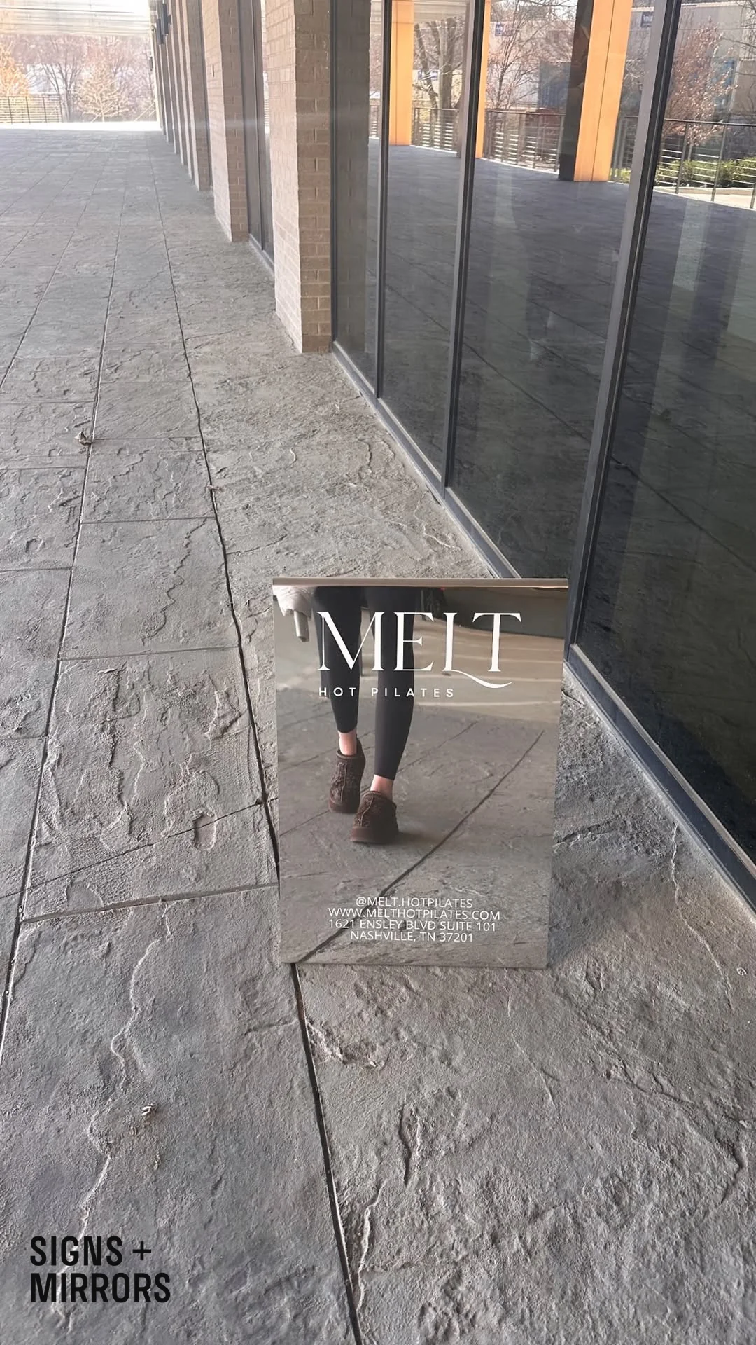 A mirrored sign with the text "MEIT HOT PILATES" and contact info is propped up outside a building with large windows. The reflection shows a person's legs and the surrounding sidewalk. "SIGNS + MIRRORS" is in the bottom-left corner.