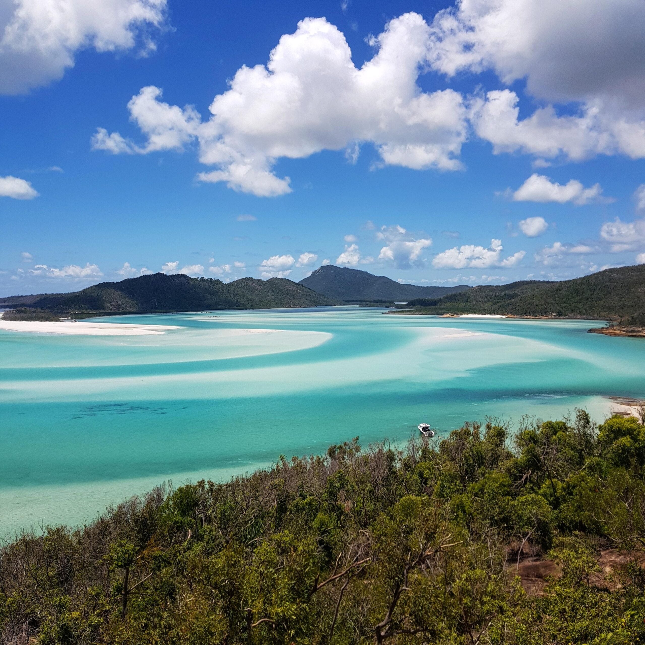 A panoramic view of a turquoise bay with swirling white sandbars, surrounded by green hills under a partly cloudy blue sky. Some foliage is visible in the foreground.