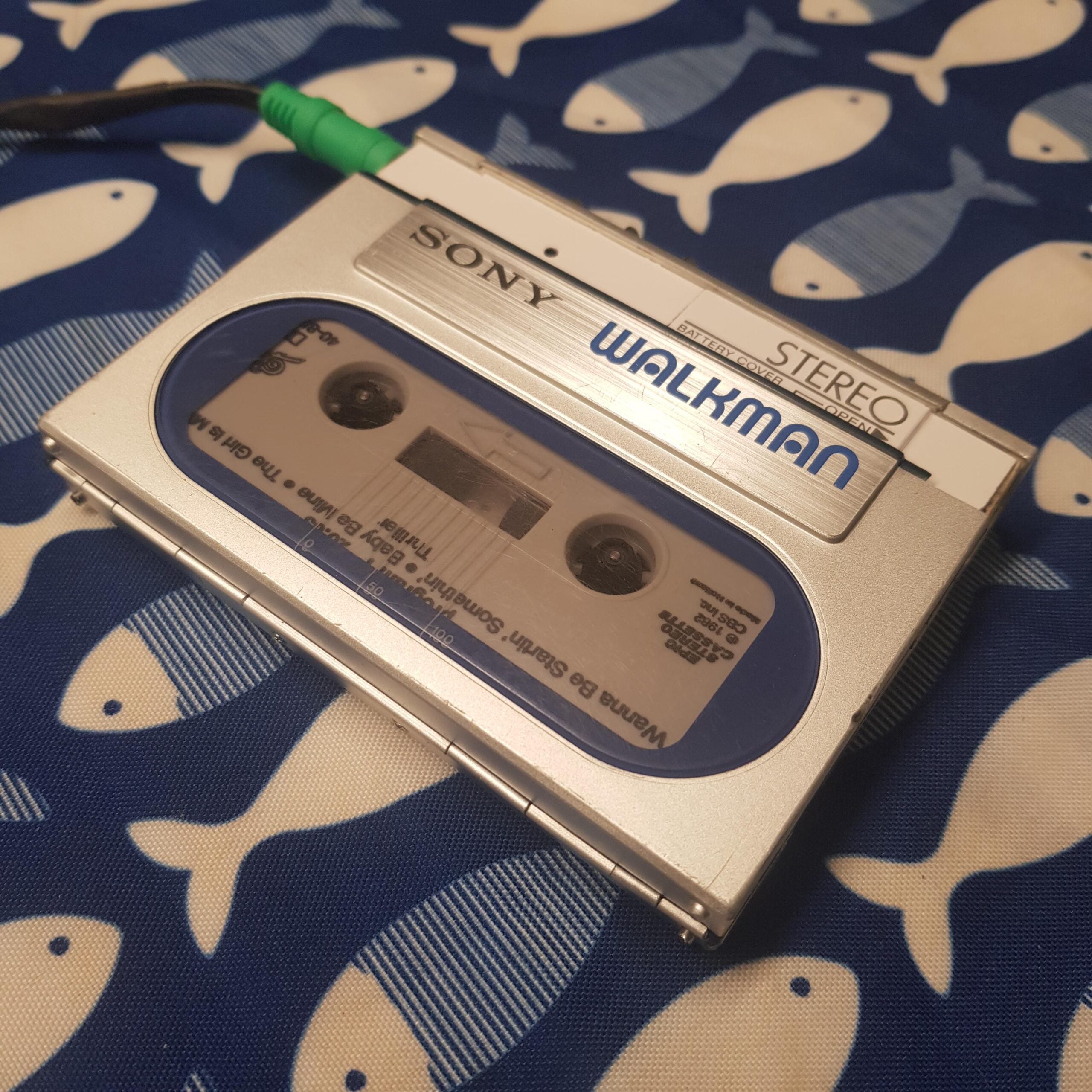 A silver Sony Walkman cassette player with a visible cassette tape inside rests on a blue and white fish-patterned fabric. A green audio jack is plugged into the Walkman.