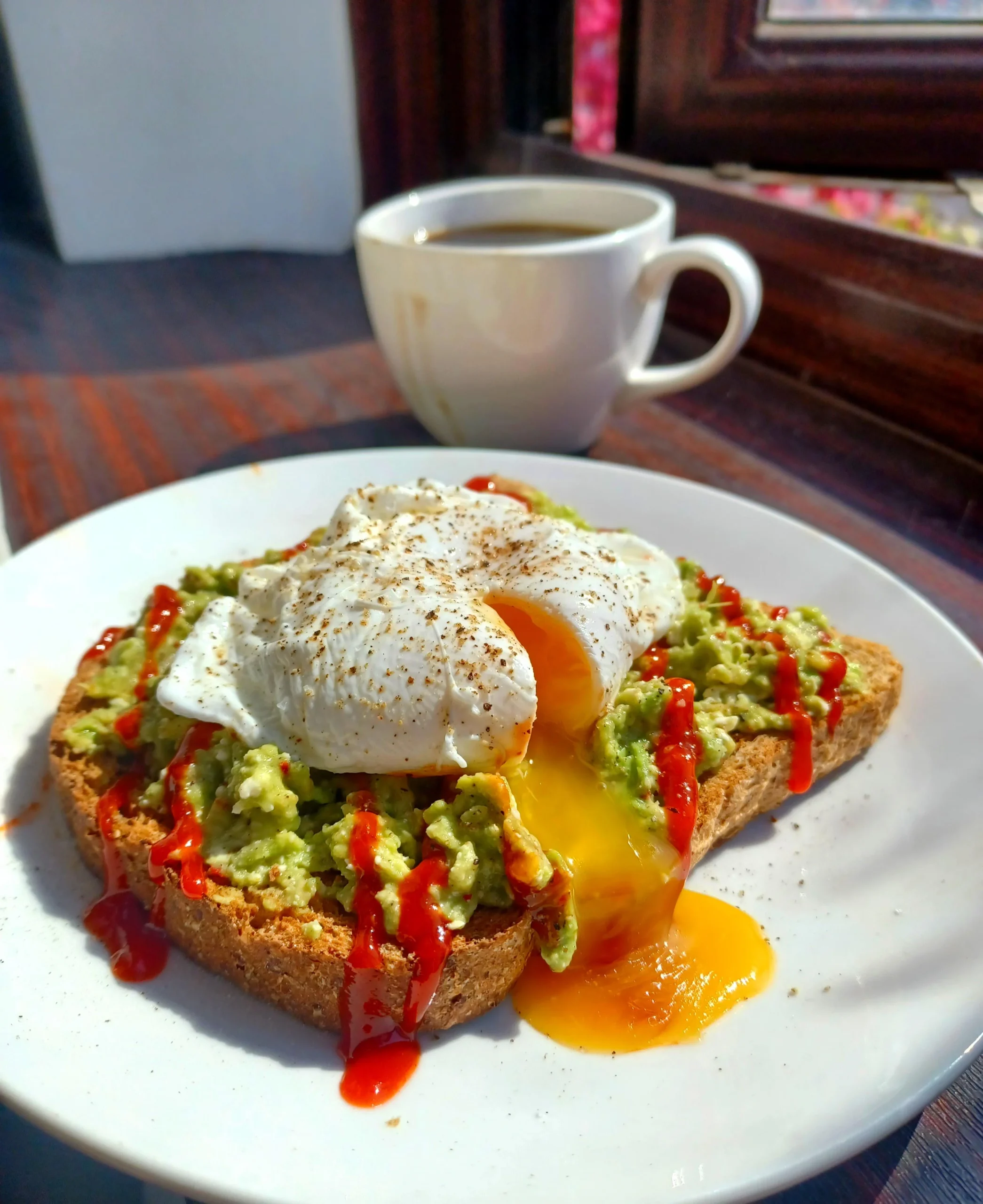 A slice of toast topped with mashed avocado, two poached eggs with runny yolks, and drizzled with hot sauce sits on a white plate. A cup of coffee is in the background on a wooden table by a window.